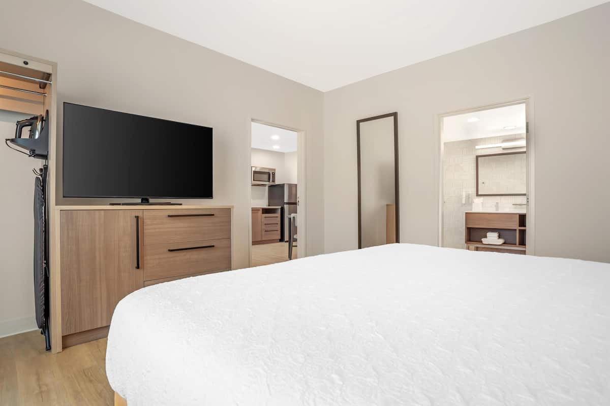 Une chambre avec un grand lit, un téléviseur posé sur une commode en bois, un miroir et, au fond, une petite cuisine.