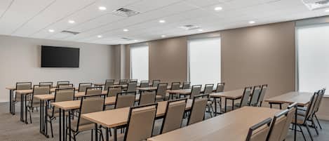 Un salón de conferencias con varias mesas y sillas, un televisor de pantalla plana montado en la pared y grandes ventanales.