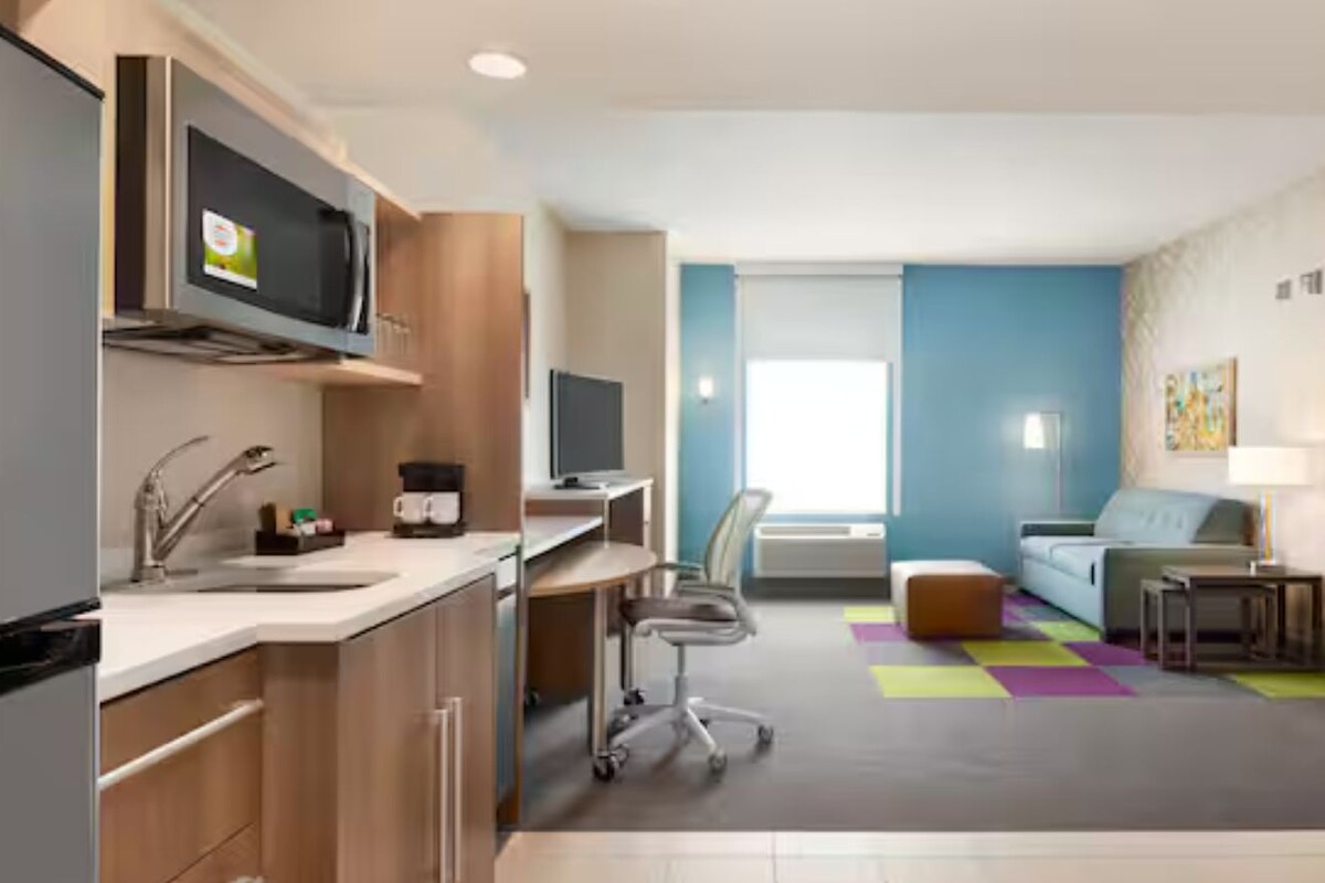 A modern hotel room with a kitchenette, a flat-screen TV, a sofa, a small table, and a colorful rug.