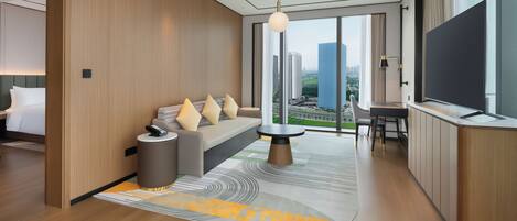 A modern hotel room with a large window, a sofa with cushions, a small table, and a TV.