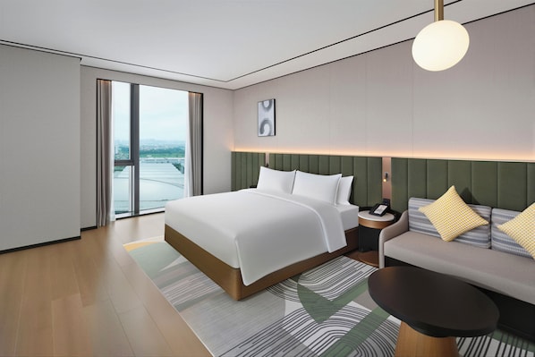 A modern hotel room with a large bed, a sofa, a round table, and a view of the city through large windows. - Hilton Garden Inn Foshan Tanzhou ICEC (Foshan)