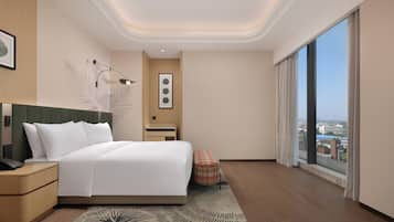 A modern hotel room with a large bed, a nightstand, a desk, a chair, a view of the city, and a large window.