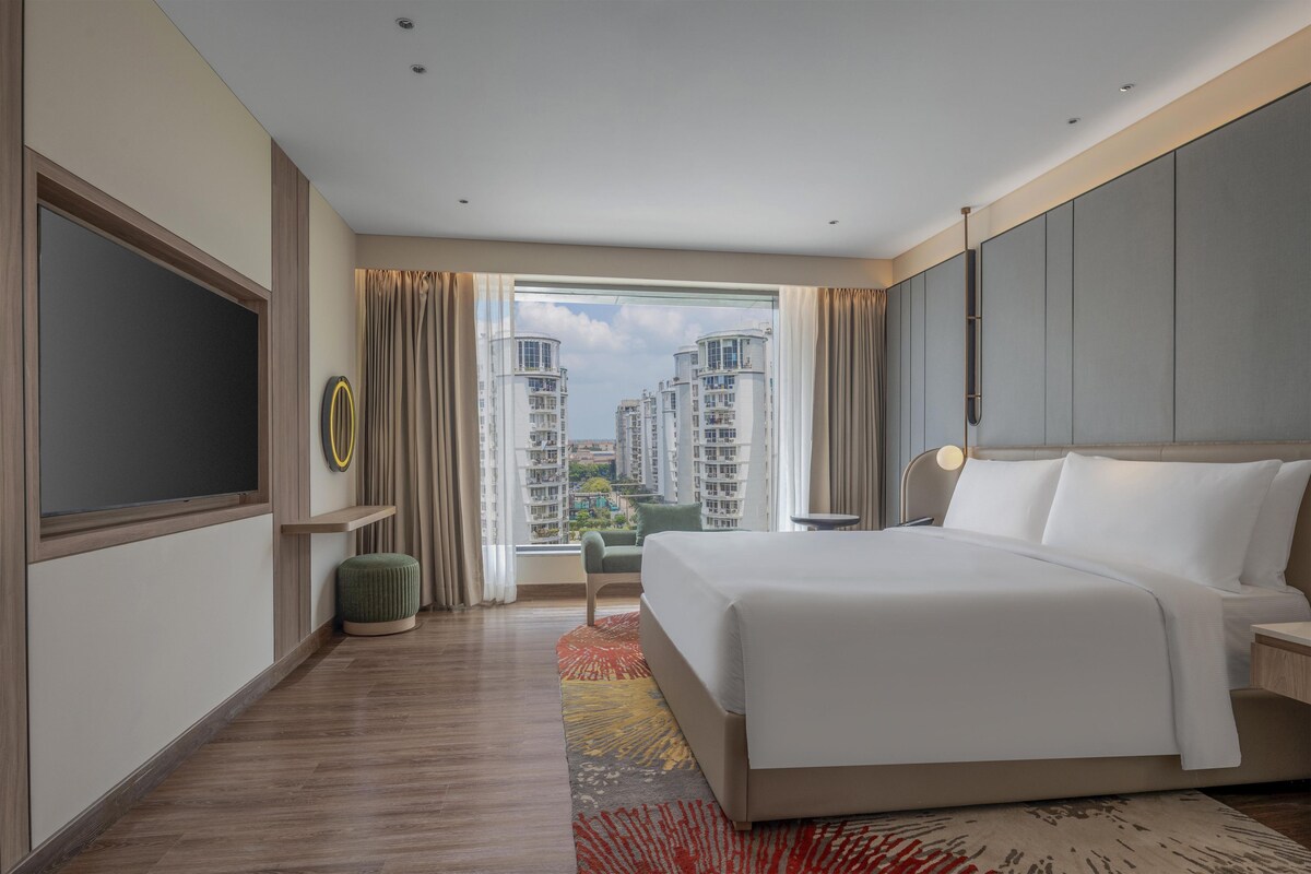 A hotel room with a large bed, a TV, a view of high-rise buildings, and a window with curtains.