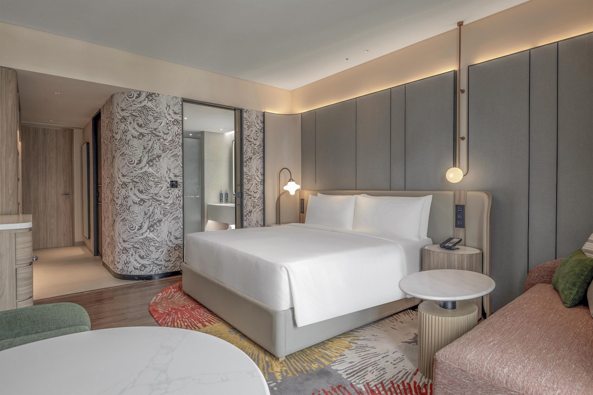A modern hotel room with a large bed, bedside tables, a seating area, and a bathroom with a mirror and towel rack.