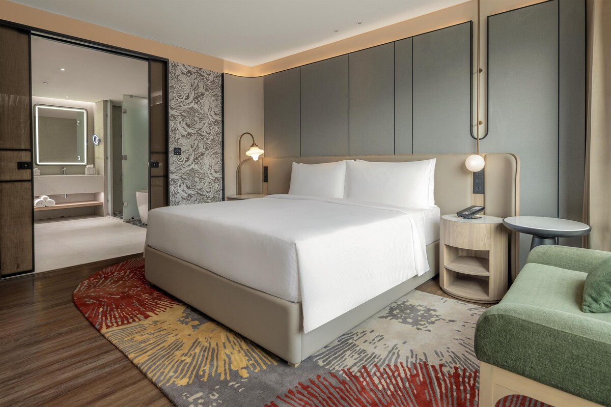 A hotel room with a large bed, bedside tables, a chair, a bathroom with a mirror, and a patterned rug.