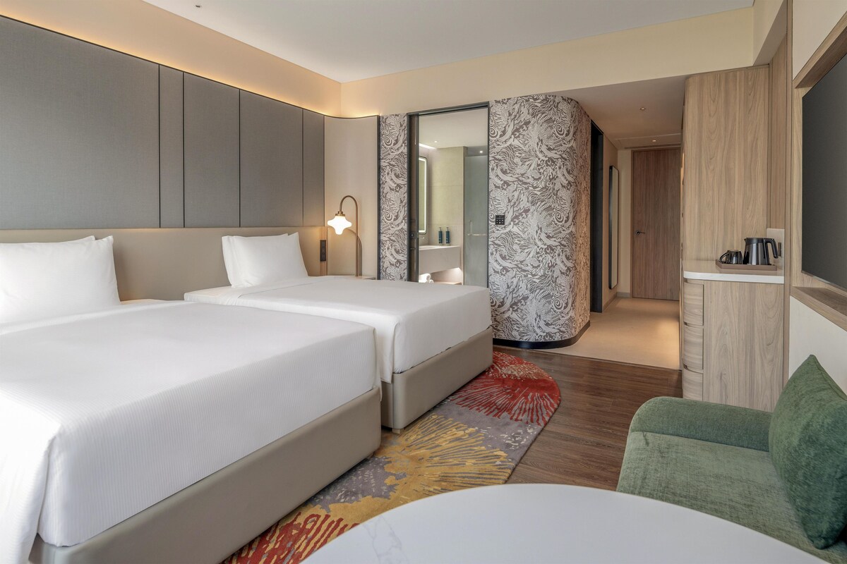 A hotel room with two beds, a sitting area with a green chair, a small table, and a shower area.