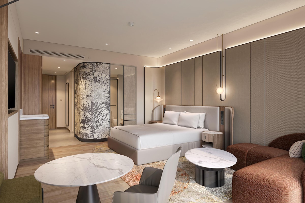 A modern hotel room with a large bed, a sitting area with a table and chairs, a mirror, and a decorative wall panel.