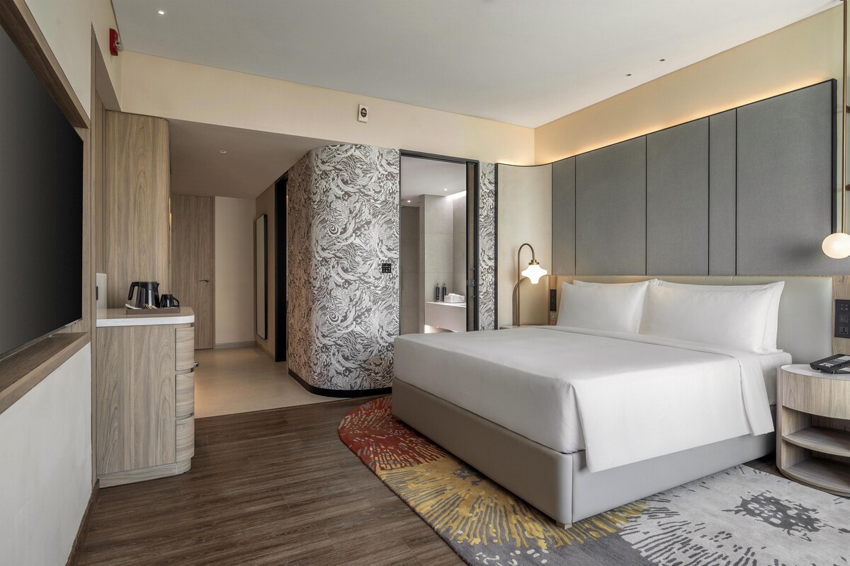 A modern hotel room with a large bed, a bedside table, a desk with a chair, a wardrobe, and a patterned wall.