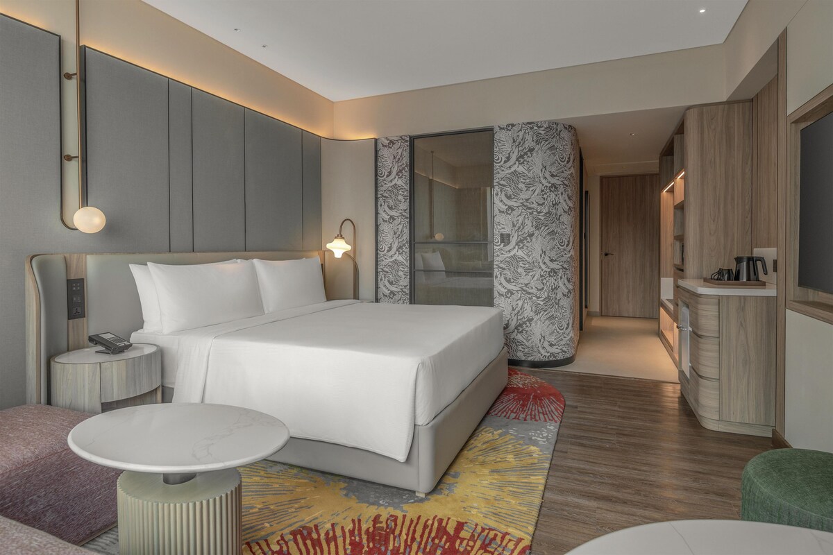 A modern hotel room with a large bed, bedside tables, a small round table, a wardrobe, and a mirror.