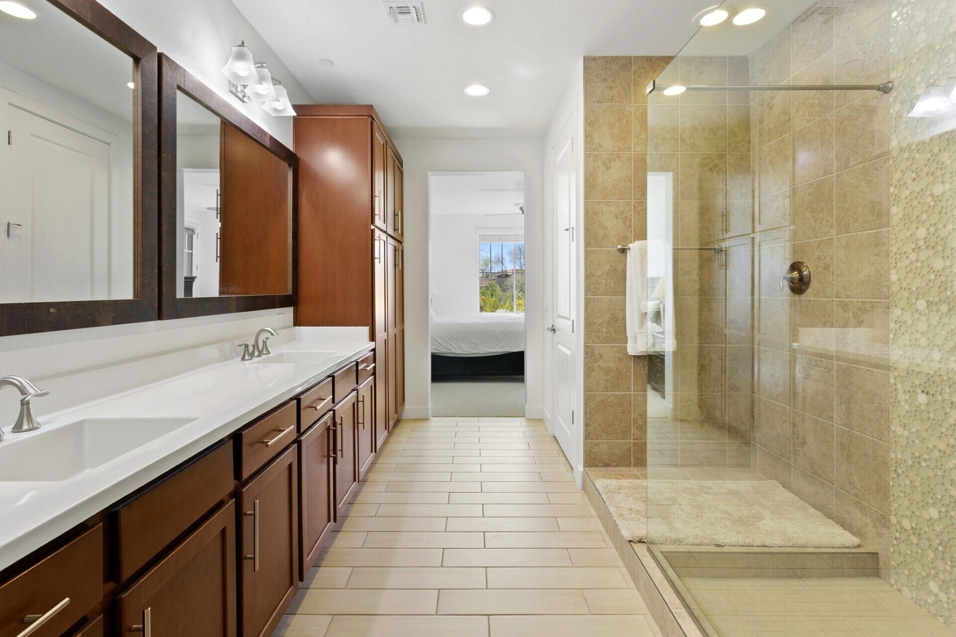 A modern bathroom with a large mirror, dual sinks, a walk-in shower, and a bed visible through an open door.
