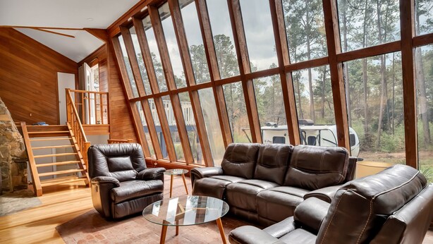 A spacious room with a large window, a leather sofa, a glass coffee table, and a wooden staircase. - Unique Durham Cabin with Hot tub and close to Duke (Durham)
