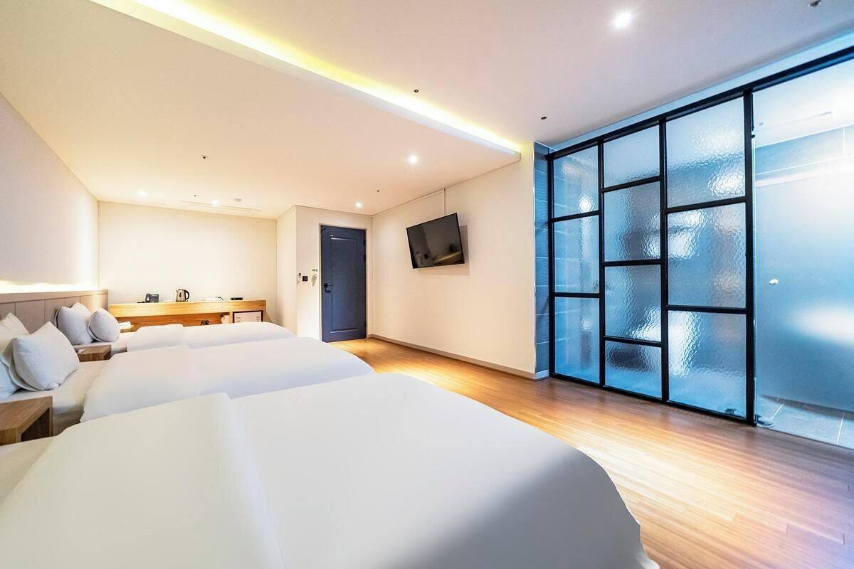 A modern hotel room with two beds, a flat-screen TV, a large window with frosted glass, and a small kitchenette.
