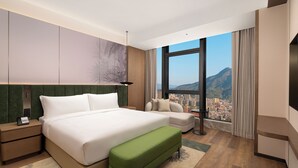 Ett modernt hotellrum med en stor säng, en grön bänk, en soffa, en tv och utsikt över staden genom stora fönster.