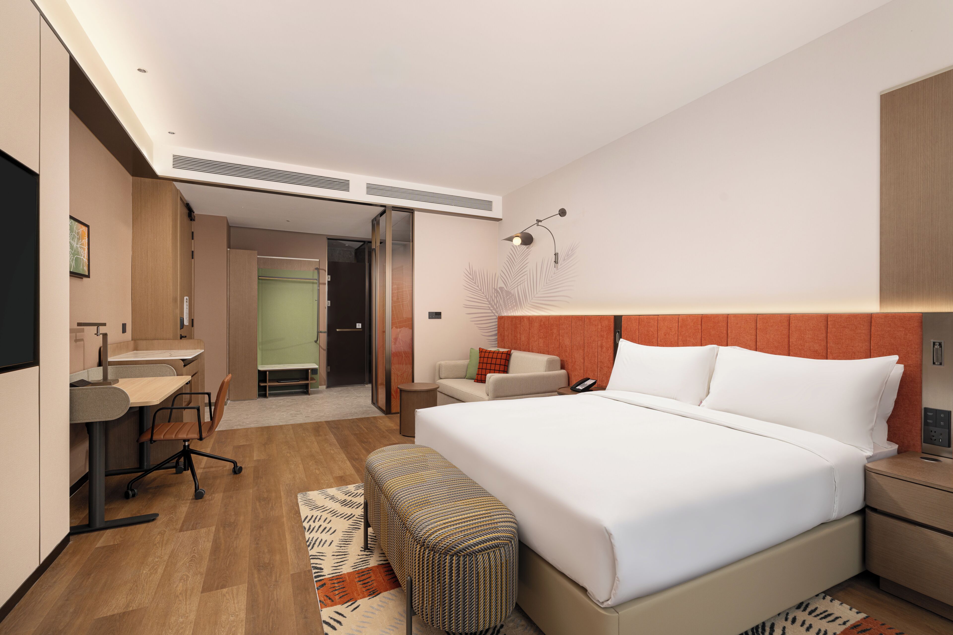 Una habitación de hotel moderna con una cama grande, un escritorio con silla, una mesita, un banco y una mesita de noche con lámpara.
