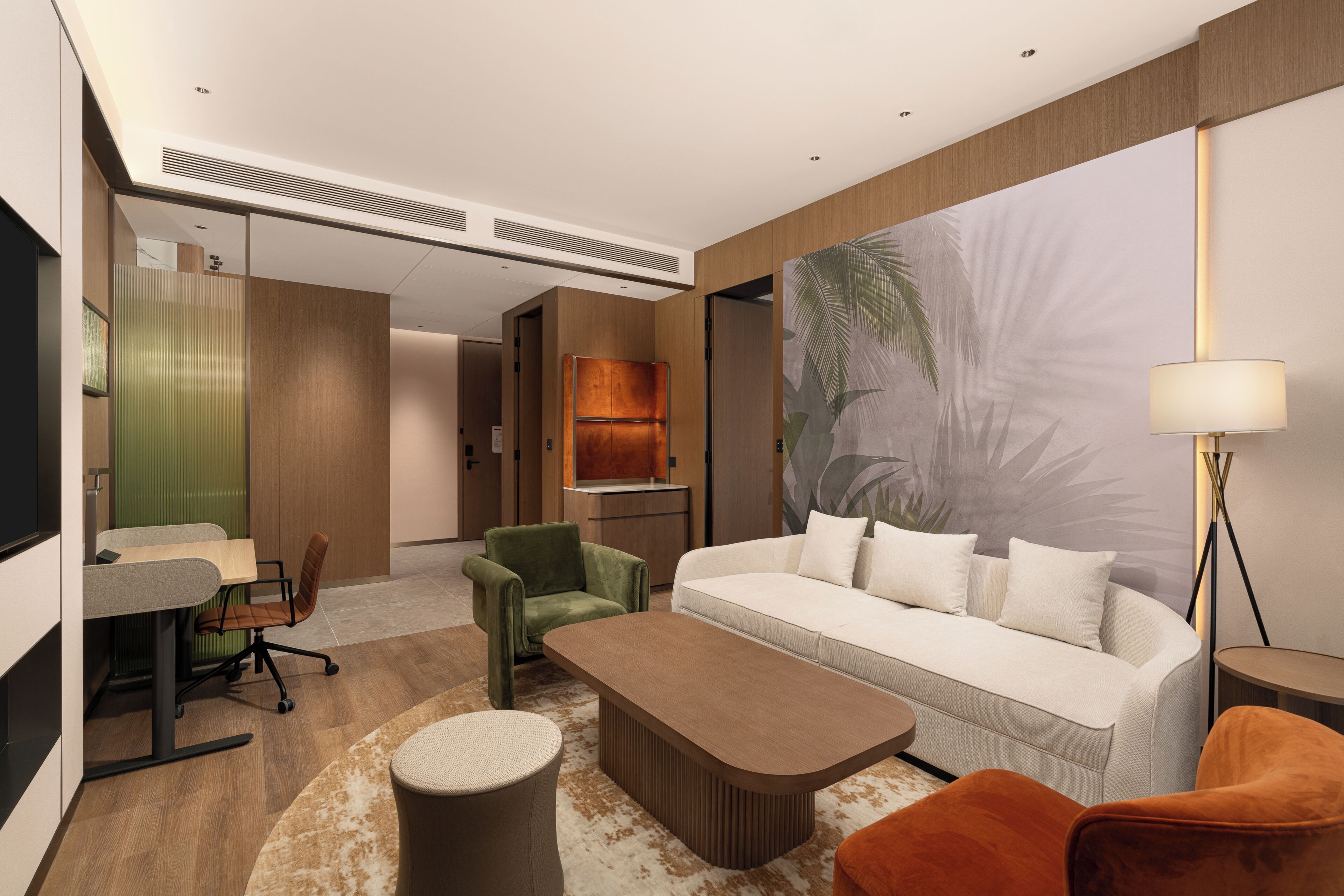 A modern hotel room with a sofa, armchair, coffee table, desk, and lamp.