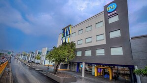A modern hotel building with a prominent 'TRU by Hilton' sign, a tree planter with a single tree, and a storefront with a purple sign. - Tru By Hilton Mexico City Airport (Mexico City)