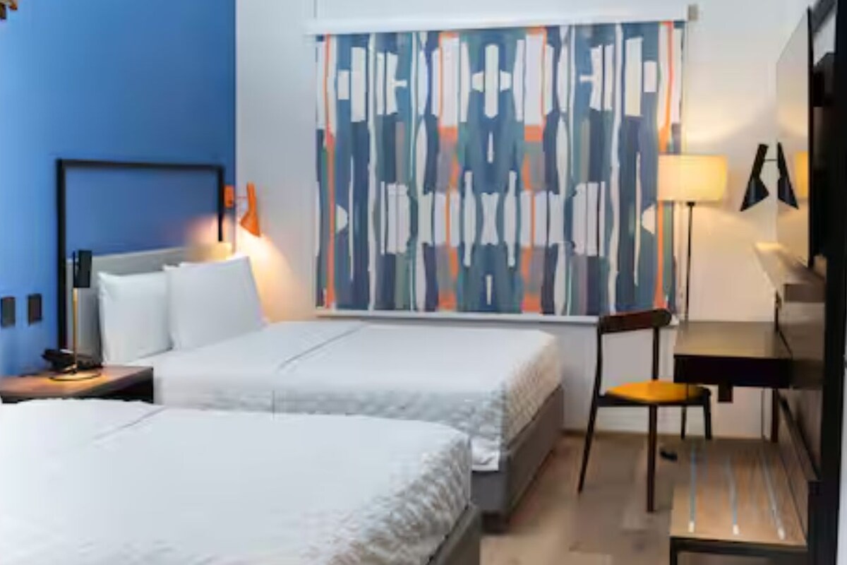 A hotel room with two beds, a desk, a chair, a lamp, and a large abstract curtain.