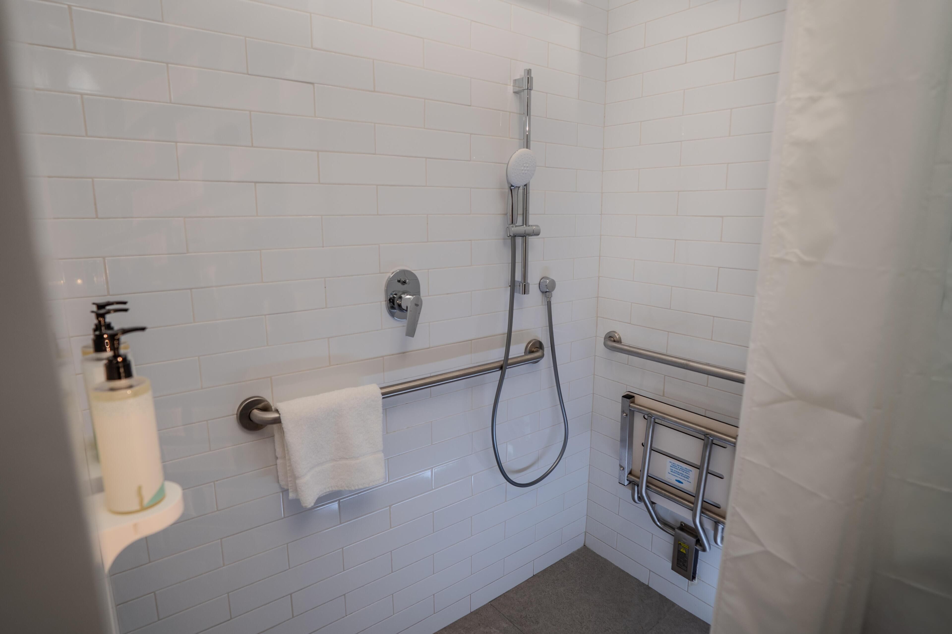 Un baño con paredes de azulejos blancos, una ducha con cabezal de mano, un perchero para toallas y un dispensador de jabón.