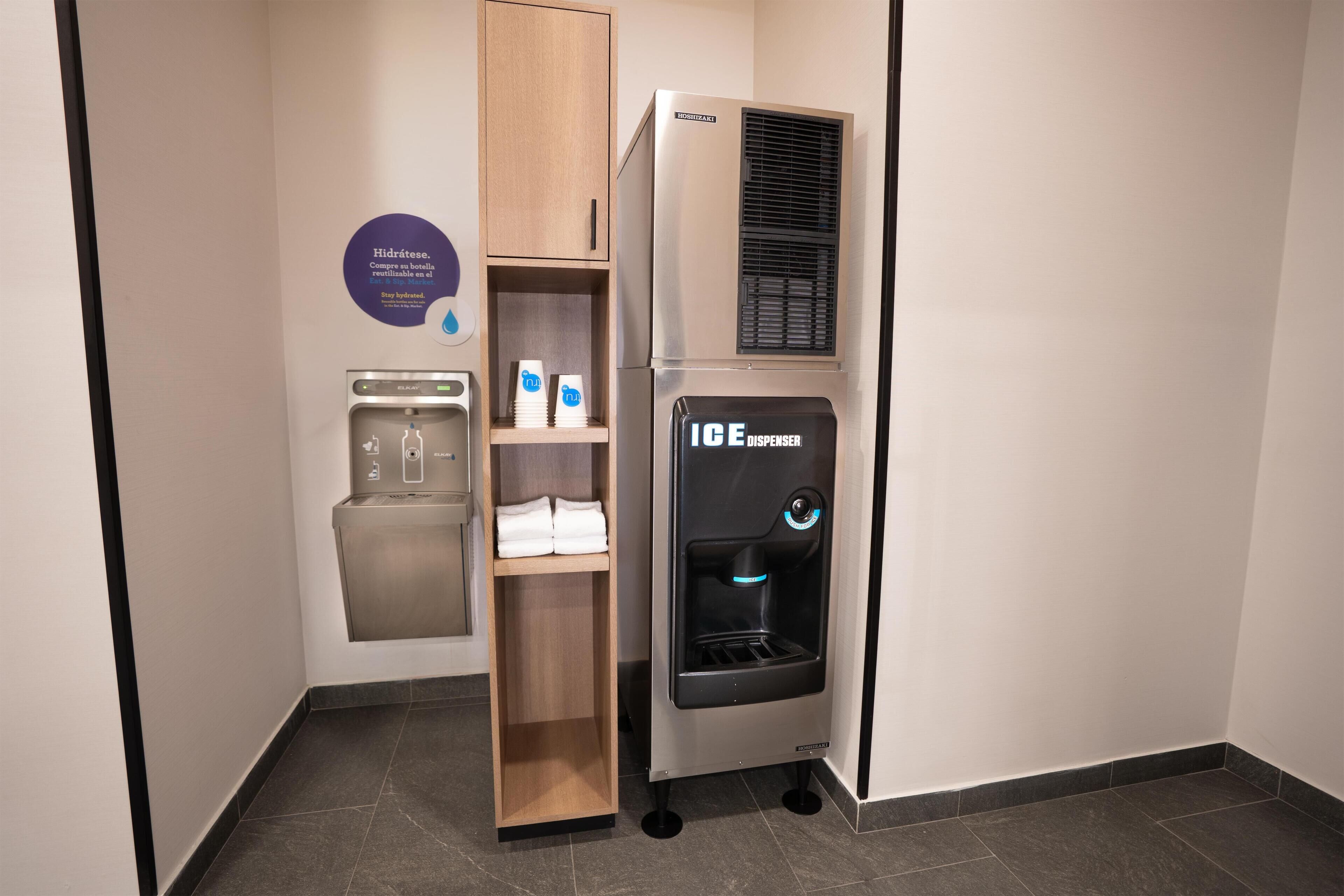 En vattenautomat, en dryckesautomat och en hylla med hopvikta handdukar i ett hotellrum.