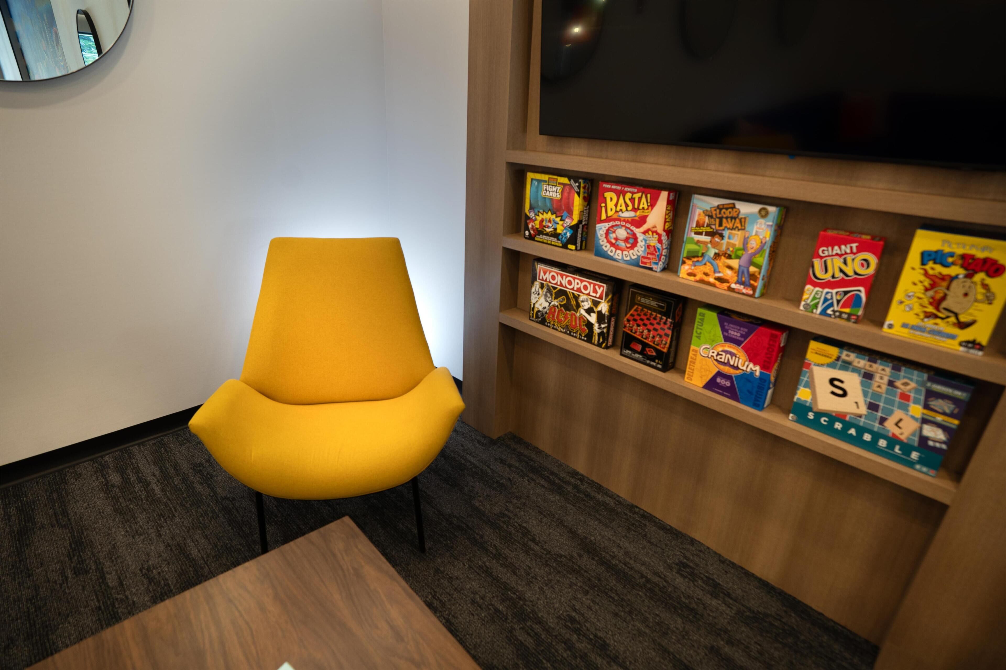 Una habitación con una silla amarilla, un estante de madera con juegos de mesa, un televisor y suelo alfombrado.