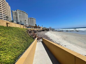 Uma passarela costeira com área ajardinada, uma praia arenosa e edifícios altos ao fundo.