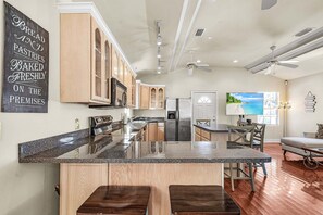 A kitchen with a granite countertop, wooden cabinets, a stainless steel refrigerator, and a dining area with a bench and table. - Steps to the Beach, Scenic Deck Views & Near Times Square Fun - FMB - The Upper Deck - Roelens (Fort Myers Beach)