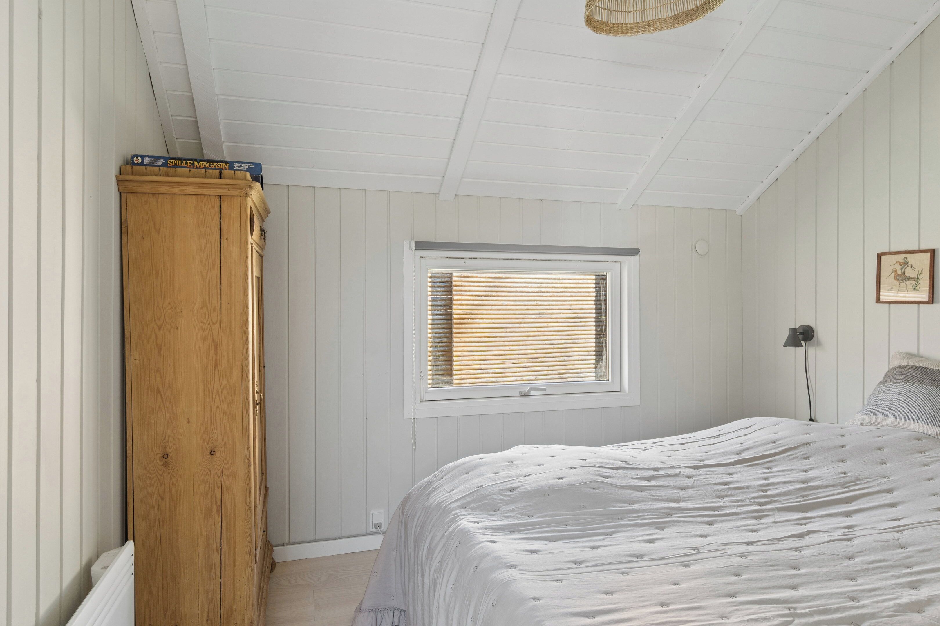 Un dormitorio con un armario de madera, una ventana con persianas, una cama con colcha blanca y una lámpara fijada a la pared.