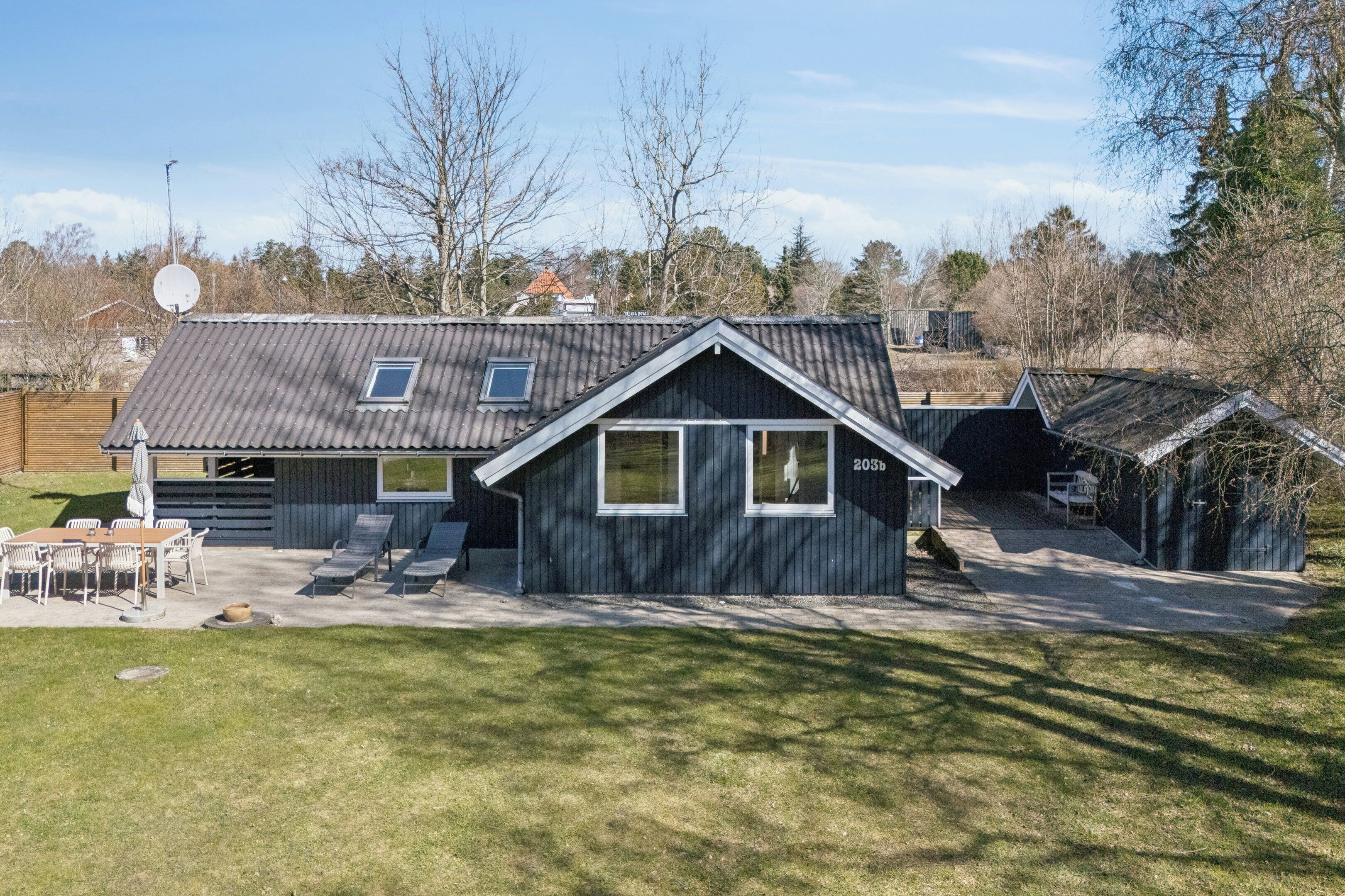 Une maison avec un toit en pente, un petit garage attenant et une terrasse aménagée avec du mobilier d’extérieur.