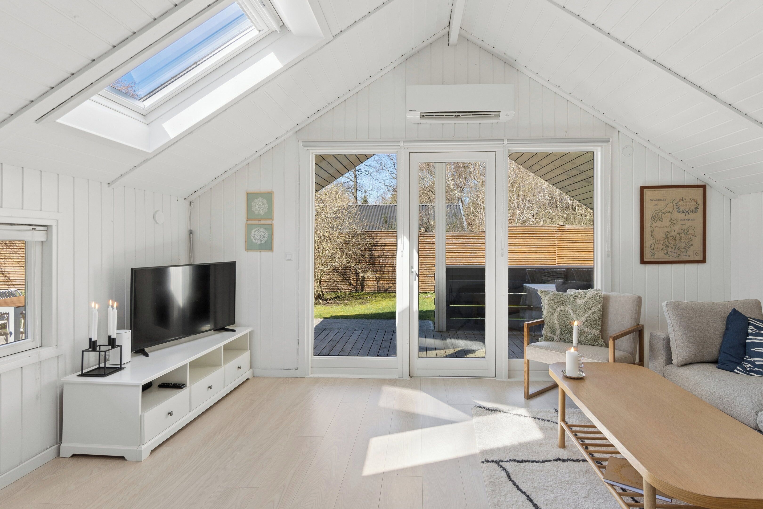 A modern living room with a TV, white furniture, a large sliding glass door to a deck, and a skylight.