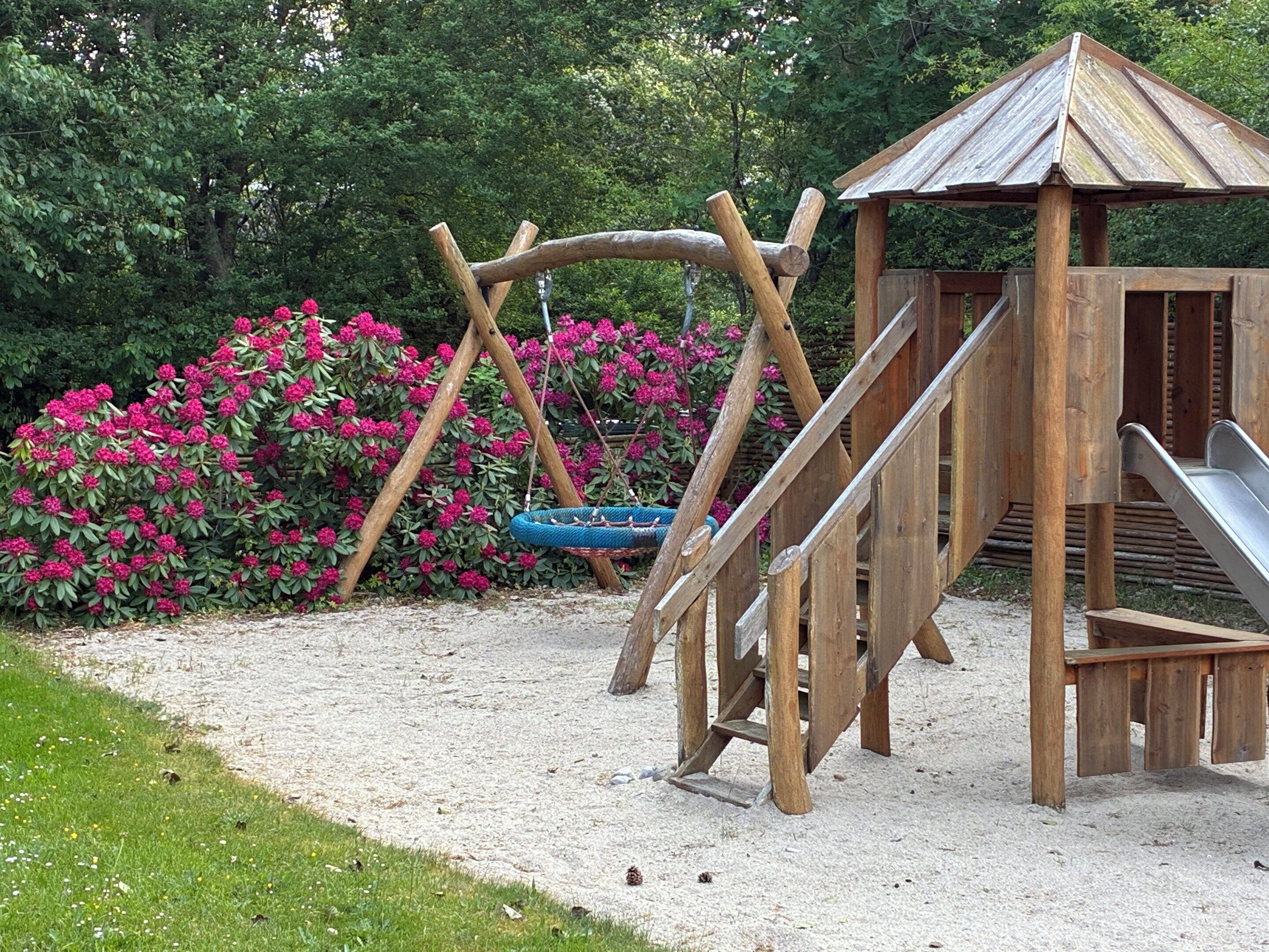 Une structure de jeux en bois, comprenant des balançoires et un toboggan, entourée de fleurs roses et de verdure.