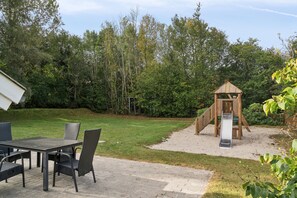 A patio with a table and chairs, a wooden play structure with a slide, and a grassy area with trees. - Lovely cottage close to a beautiful beach (Vig)