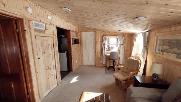 Et interiør af en træhytte med en sofa, en stol, et lille bord, et skrivebord med stol og et vindue med gardiner.