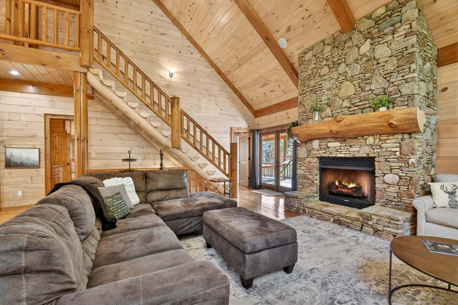 A log cabin interior with a stone fireplace, a wooden staircase, and a sectional sofa.