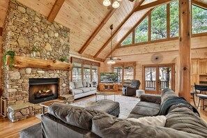 A log cabin interior with a stone fireplace, wooden beams, and large windows. - Kick Aska Lodge・Hot Tub・Firepit・Game Room・Secluded (Cherry Log)