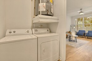 Un espace buanderie équipé d’un lave-linge et d’un sèche-linge, une petite table et un canapé bleu, le tout dans une pièce au plancher en bois et dotée d’une fenêtre avec des stores.