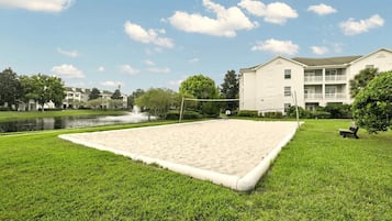 Ein Sandvolleyballfeld, umgeben von Gras, mit Teich und Bäumen im Hintergrund.