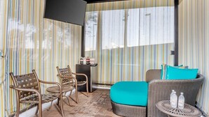 Una habitación con cortinas a rayas, una cama con cojín color turquesa, muebles de mimbre y una pequeña mesa con botellas.