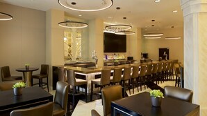 Un área de bar moderna con diversas opciones de asiento, incluyendo taburetes y mesas, y un televisor de pantalla plana grande montado en la pared.