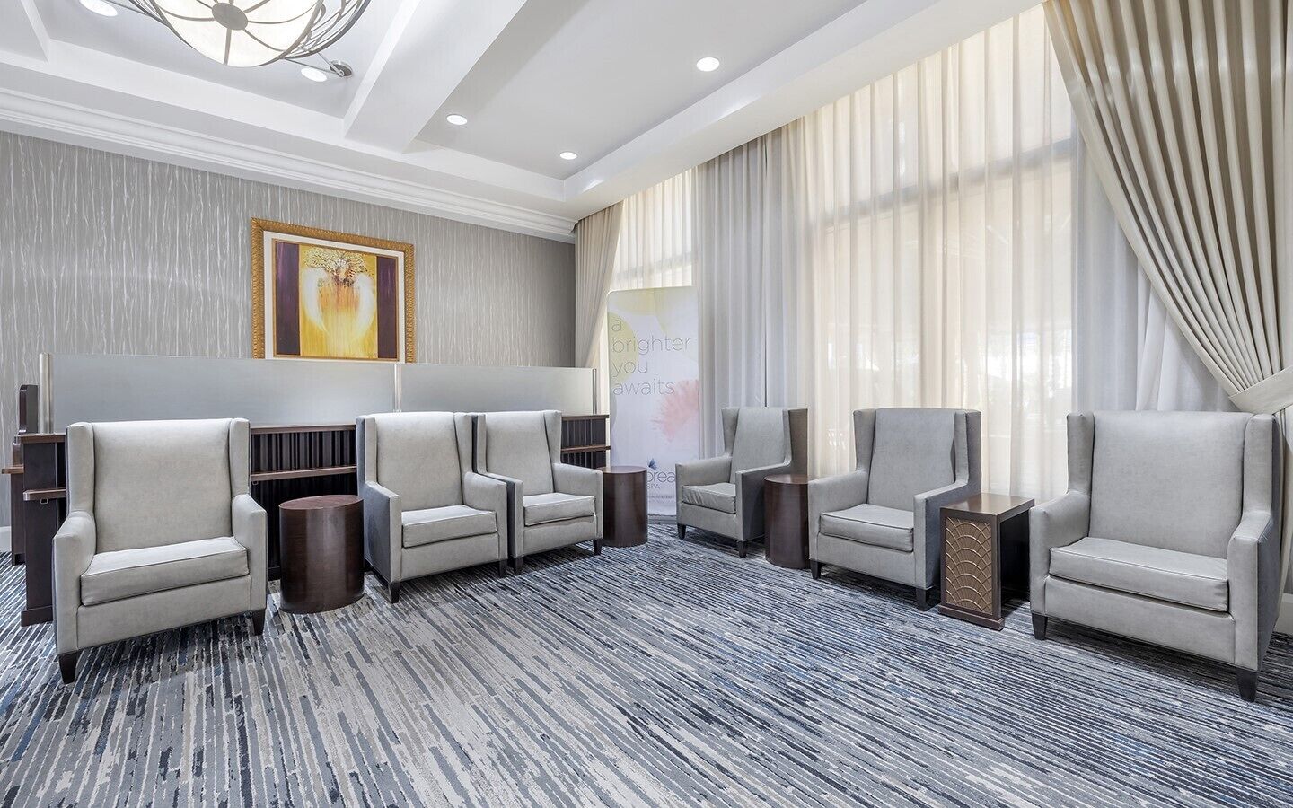 A hotel lobby with a seating area, a large window with curtains, a painting on the wall, and a carpeted floor.