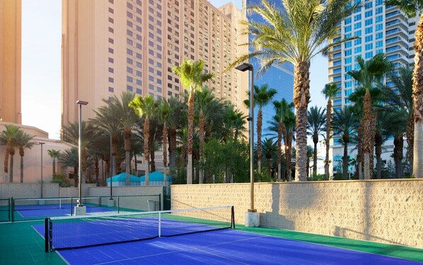 A tennis court with a net, surrounded by palm trees and high-rise buildings.