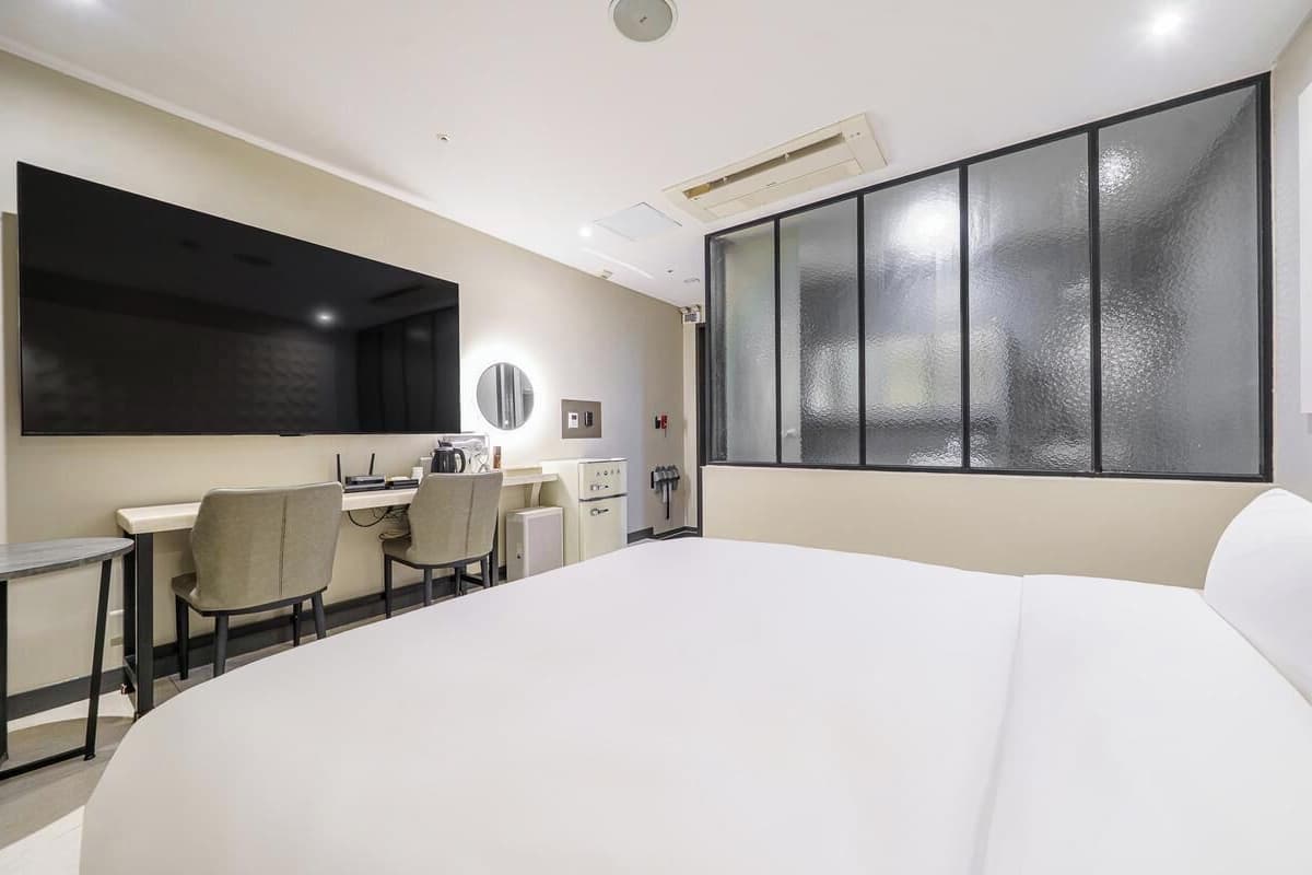 Habitación de hotel con una cama grande, un escritorio con sillas, un televisor de pantalla plana, un ventanal con vidrio esmerilado y un equipo de aire acondicionado instalado en la pared.
