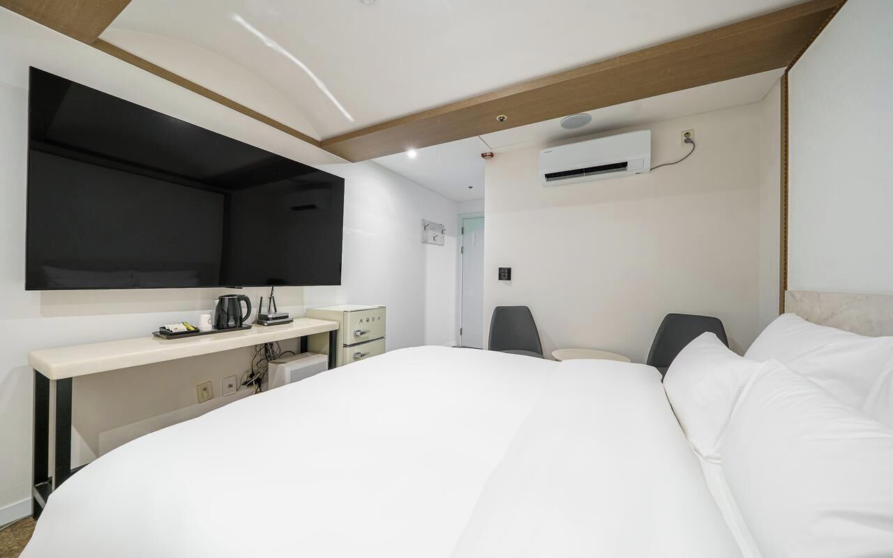 A modern hotel room with a large bed, a desk with a kettle, a chair, and an air conditioning unit.