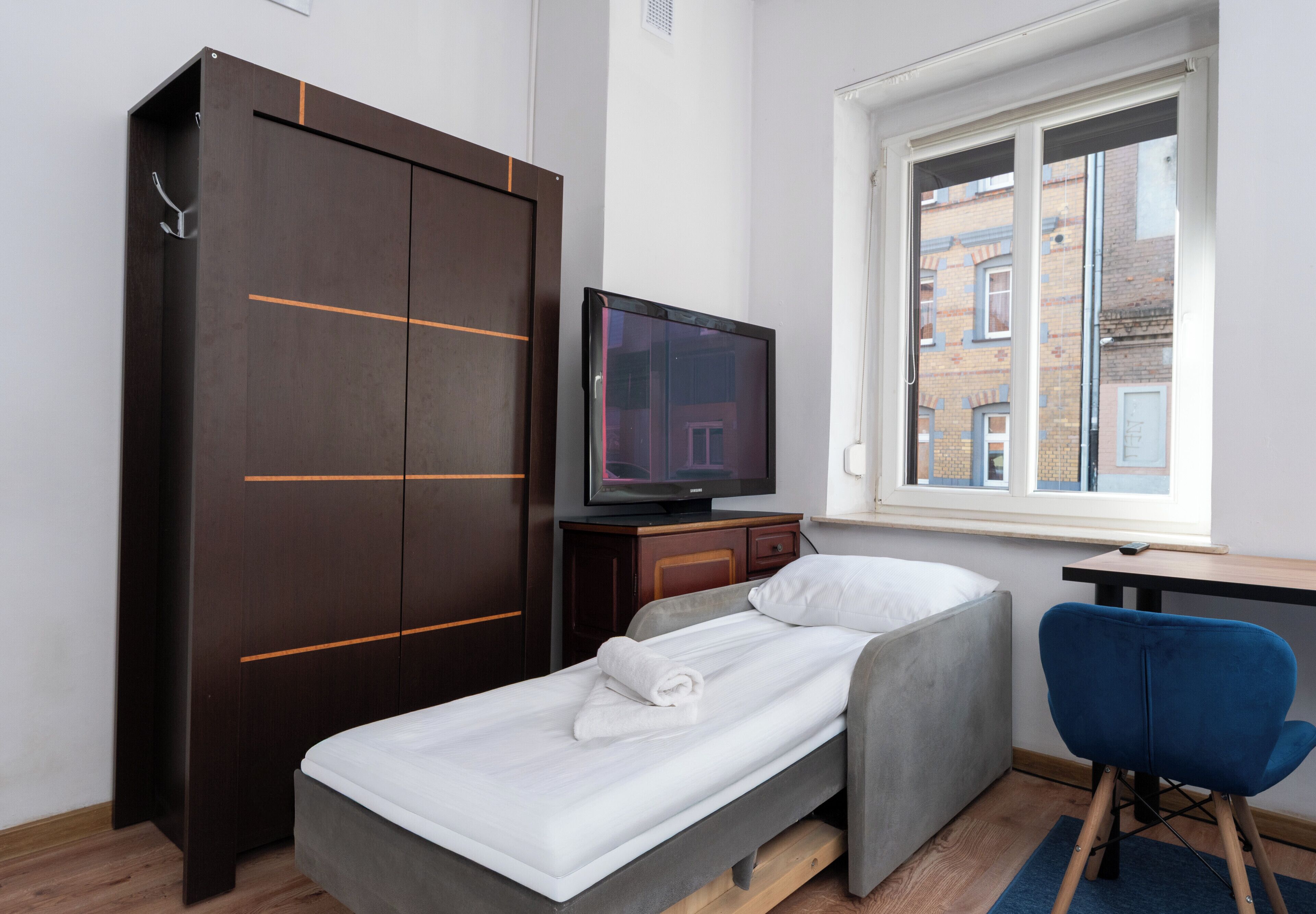 A single bed with a white mattress and grey bedding, a television on a wooden stand, a blue chair, and a large wardrobe with horizontal lines.