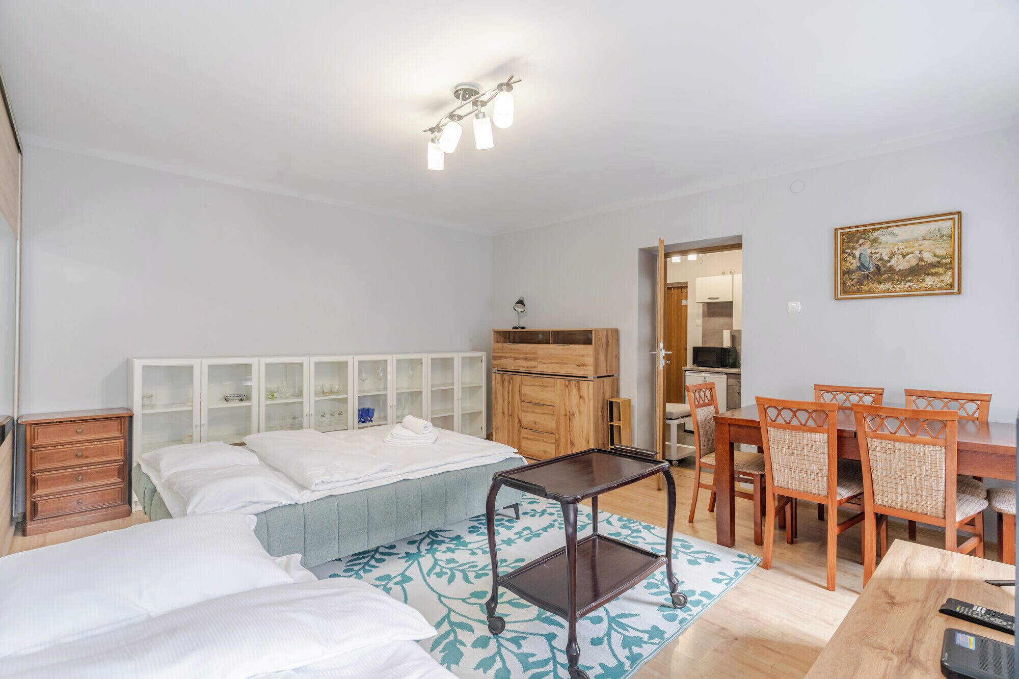 Une chambre avec deux lits, une armoire en bois, une table à manger avec des chaises, et un coin cuisine à l’arrière-plan.