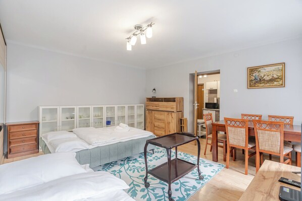 Ein Schlafzimmer mit zwei Betten, einem Holzschrank, einem Esstisch mit Stühlen und einer Küchenzeile im Hintergrund.