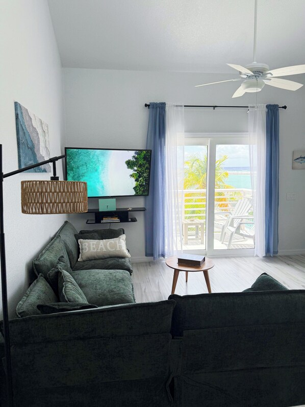 Una sala de estar con un sofá, un televisor, un ventilador de techo y vistas al exterior.
