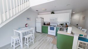 Cocina compacta con armarios blancos, encimera verde, refrigerador blanco, estufa y microondas. Hay una pequeña mesa de comedor con sillas blancas.