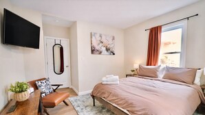 Quarto de hotel com cama, escrivaninha com cadeira, televisão, porta e janela com cortinas.