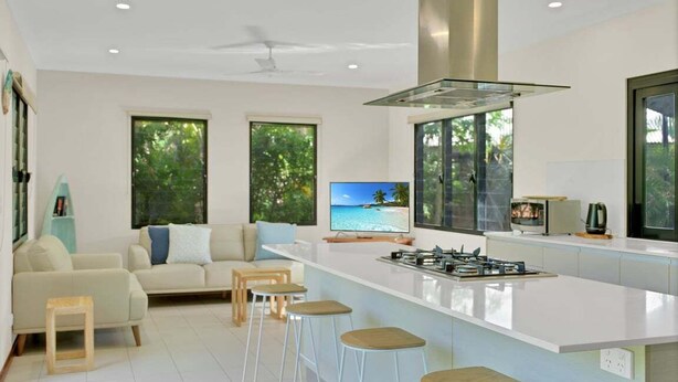 A modern kitchen with a central island, bar stools, a sofa, and a television displaying a beach scene. - Balu luxe (Broome)