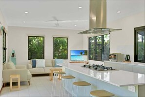 A modern kitchen with a central island, bar stools, a sofa, and a television displaying a beach scene.